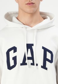 Sweat-shirt à capuche blanc avec un logo "GAP" bleu marine. Texture douce, cordons de serrage sur la capuche, poignets côtelés et coupe décontractée. Design simple.