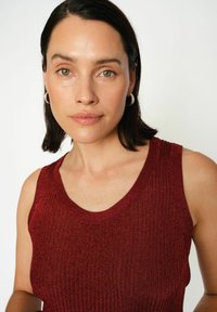 Geribbeld rood mouwloos topje met een ronde halslijn, voorzien van een gemêleerd breipatroon. Model toont een soepele, natuurlijke make-uplook.