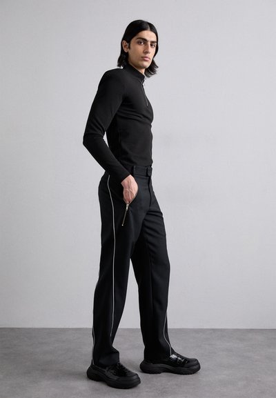 Chemise noire à manches longues avec un col montant et une fermeture éclair sur le devant, associée à un pantalon noir avec des passepoils blancs et des poches zippées.