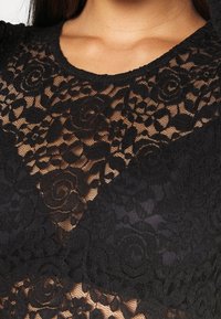 Haut en dentelle noire présentant des motifs floraux complexes, une texture transparente et un col rond, mettant en valeur un design délicat et superposé.