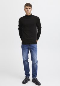 Maglione nero a collo alto con maniche lunghe, abbinato a jeans in denim blu. La modella indossa scarpe nere senza lacci, in piedi su uno sfondo uniforme.