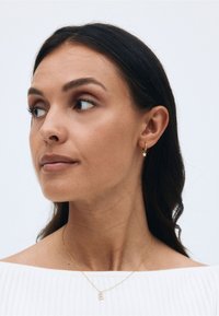 Guldstjärnformad hoopsring, delikat kedjesmycke med 'E' hänge, modell som bär en off-shoulder vit texturerad topp, neutral bakgrund.