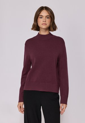 Pull marron à col montant et manches longues. Présente une texture douce et un ourlet côtelé. Associé avec un pantalon noir côtelé.