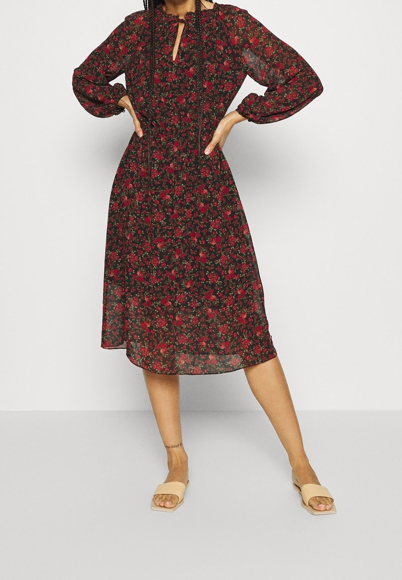Femme portant une robe midi noire à imprimé floral rouge, manches longues, et des sandales béiges, debout les mains sur les hanches.
