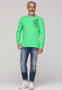 Maglietta a maniche lunghe verde con dettagli testuali grafici neri, abbinata a jeans azzurri chiari e sneakers bianche su uno sfondo neutro.