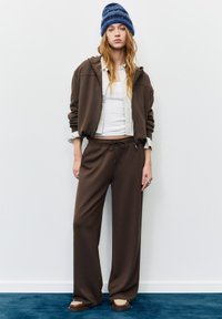 Smeđa majica s kapuljačom i usklađene trenirke izrađene od mekane tkanine, s bijelom majicom ispod i plavom prugastom kapom. Obuća ima bež naglasak.