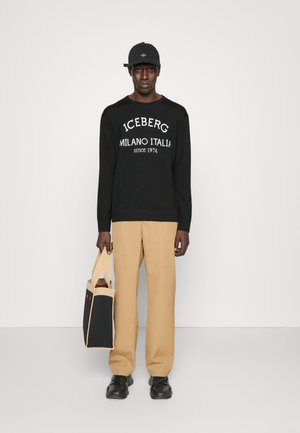 Un pull noir avec le texte blanc "ICEBERG MILANO ITALIA", un pantalon beige, des chaussures noires et une casquette noire tenant un sac fourre-tout bicolore.