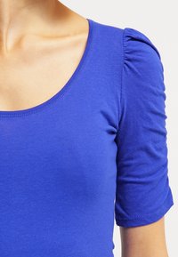 Top azul de manga corta con escote redondo, que presenta detalles de tela gather en los hombros y una textura suave y elástica.