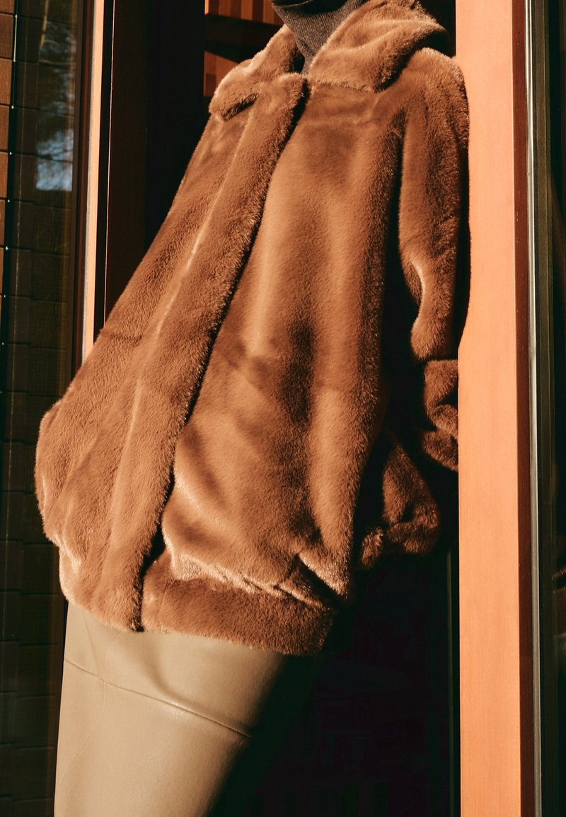 Hnědá bunda z umělé kožešiny s límcem, nadměrný střih, texturovaný povrch a spadená ramena, kombinovaná s hladkou, přiléhavou béžovou sukní.