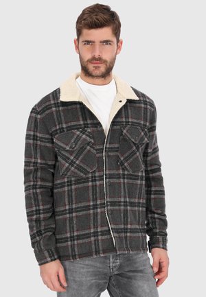 Mann trägt eine grau-rot karierte Jacke mit weißem Fleecefutter und zwei Brusttaschen über einem weißen Hemd und grauen Jeans.