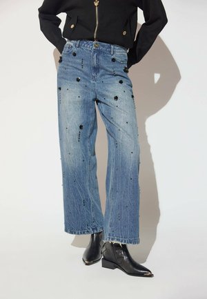 Jean bleu ample taille haute avec des ornements en perles noires, porté avec des bottines noires et une veste noire zippée.