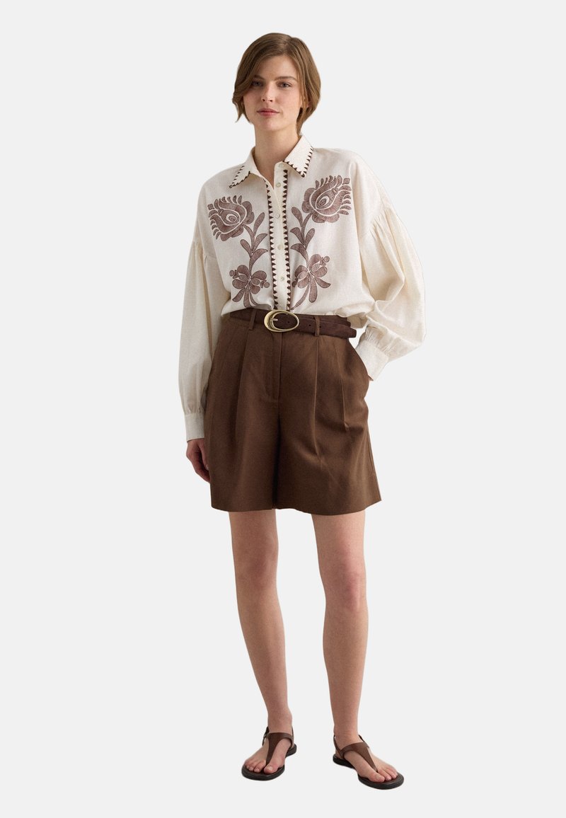 Mujer con blusa crema con bordado floral marrón, shorts marrones con cinturón y sandalias marrones, de pie contra un fondo liso.