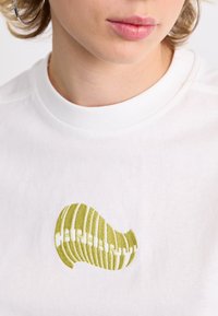 White cotton t-shirt featuring a ribbed green fishbone design embroidered on the front. Fabric is smooth with a crew neckline.