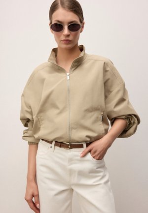 Vrouw met beige jas met rits en opgerolde mouwen, witte broek, bruine riem en donkere ovale zonnebril, staand tegen een effen achtergrond.