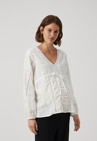 Hvit bluse med lange ermer, v-hals, knyting i midjen og åpen strikkedesign på ermene, laget av teksturert stoff.