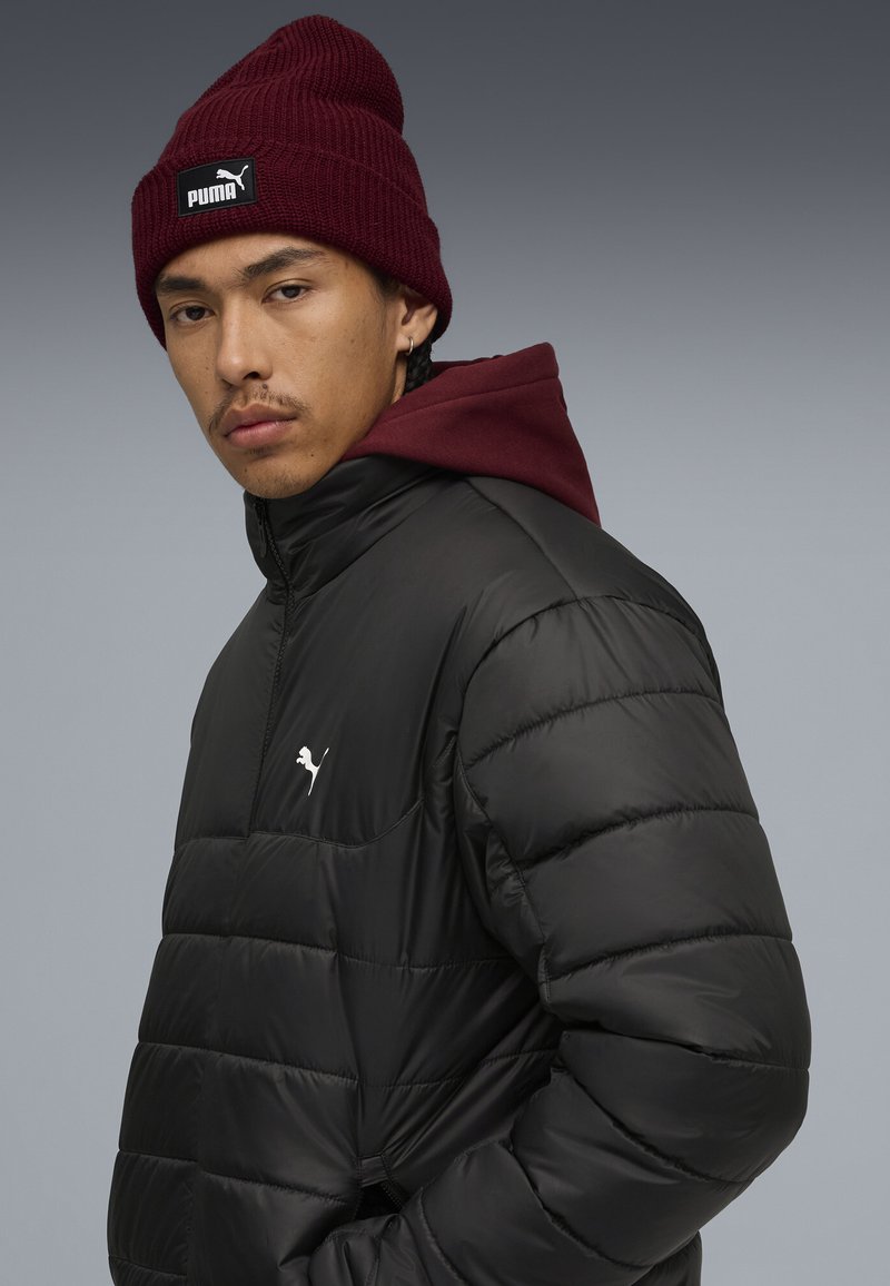 Giacca a vento nera con trama trapuntata, collo alto e logo Puma bianco. Abbinata a un berretto lavorato a maglia bordeaux con etichetta nera.