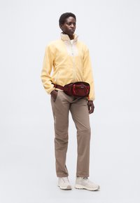 Άτομο που φοράει κίτρινο φλις μπουφάν, μπεζ παντελόνια, λευκά αθλητικά παπούτσια και κόκκινη τσάντα μέσης, στέκεται με τα χέρια στις τσέπες σε λευκό φόντο.