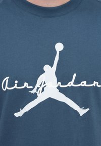 Μπλε βαμβακερό t-shirt με μεγάλο λευκό λογότυπο Air Jordan που απεικονίζει έναν παίκτη να πηδάει για να σουτάρει καλαθόσφαιρα, με το όνομα της μάρκας από κάτω.