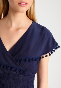 Námořnická modrá blůza s křížovým designem, má krátké rukávy, texturované smocking a dekorativní třásně podél okraje.