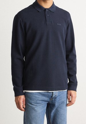 Polo à manches longues bleu marine en tissu côtelé, avec un col à deux boutons, un accent logo discret et une coupe décontractée. Porté sur un jean clair.