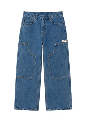 Mann mit lila gestrickter Mütze, schwarzer gesteppter Weste über einem weißen Pullover, lockeren blauen Jeans und beigen Schuhen, steht vor weißem Hintergrund.