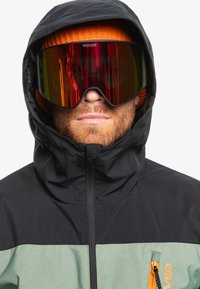 Chaqueta impermeable negra y verde con un bolsillo con cremallera, combinada con un gorro naranja y gafas de gran tamaño con lentes ahumados.