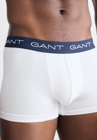 Boxer ajustados de algodón blanco con una cinturilla elástica azul marino que presenta el nombre de la marca "GANT" en blanco. Textura suave y diseño ajustado.