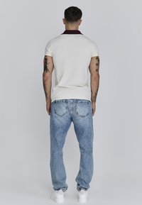 Witte polo met een kastanjebruine kraag, gecombineerd met lichtblauwe denim jeans en witte sneakers, die een relaxte pasvorm en zichtbare details op de achterkant laten zien.