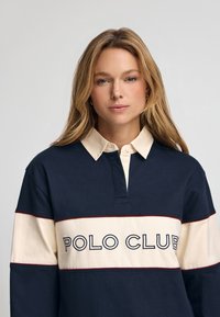 Polo a maniche lunghe in blu navy e crema, con colletto a costine, patta con bottoni e testo "POLO CLUB" in grassetto blu navy.