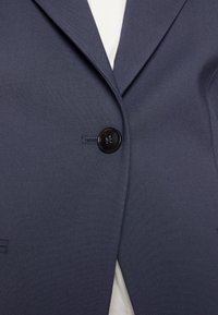 Giacca blazer blu scuro abbottonata con un singolo bottone nero su una camicia di colore chiaro.