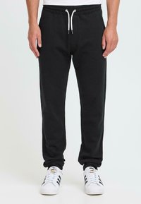 Mann in schwarzen Sweatpants mit Kordelzug und weißen Sneakers mit schwarzen Streifen, der vor einem schlichten, hellen Hintergrund steht.