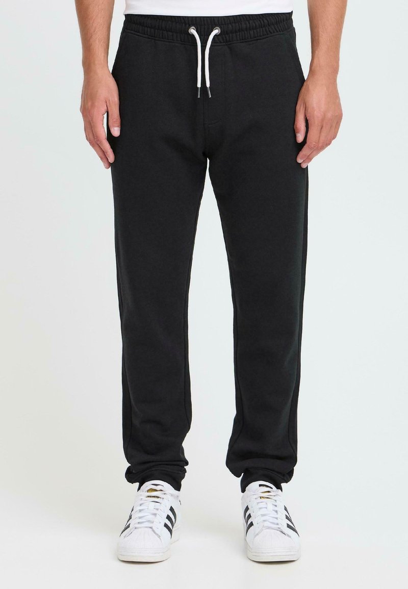 Mann in schwarzen Sweatpants mit Kordelzug und weißen Sneakers mit schwarzen Streifen, der vor einem schlichten, hellen Hintergrund steht.