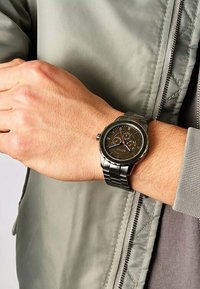 Metallische Uhr mit einem runden schwarzen Ziffernblatt, silbernen Zeigern und Zusatzanzeigen. Sie verfügt über ein silbernes Metallarmband und eine strukturierte Oberfläche.