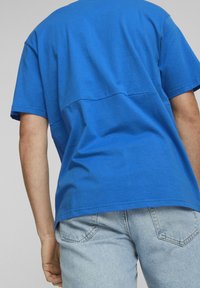 Modrá bavlněná tričko s krátkými rukávy, které má hladkou texturu a horizontální šev detail vzadu. Ležérní styl kombinovaný s lehkými džíny.