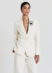 Blazer bianco su misura con maniche lunghe e scollo a V profondo, con ricamo di un fiore nero sul bavero. Tessuto liscio, vita cintata.