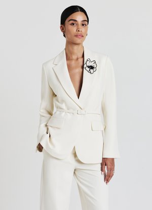 White tailored blazer with long sleeves and a deep V-neck, featuring a black flower embroidery on the lapel. Smooth fabric, belted waist.