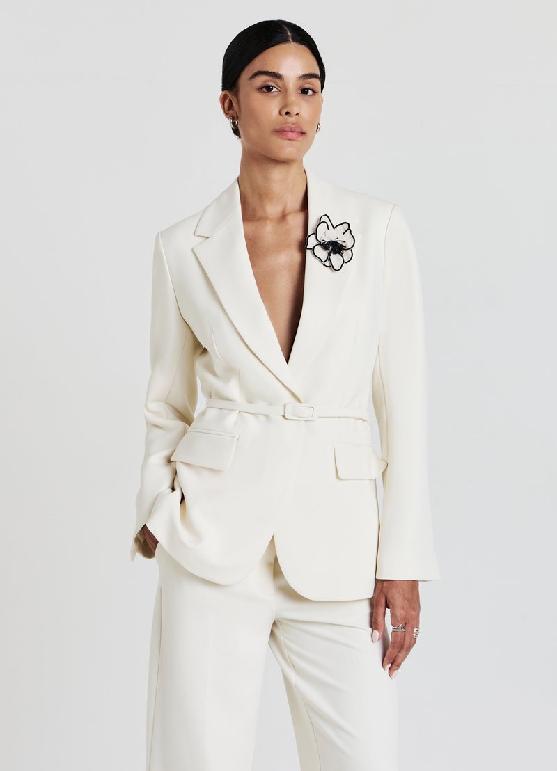 Blazer bianco su misura con maniche lunghe e scollo a V profondo, con ricamo di un fiore nero sul bavero. Tessuto liscio, vita cintata.