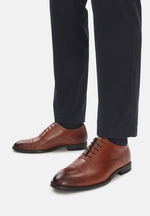 Zapatos de vestir de cuero marrón con textura suave, punta redondeada y cinco ojales para cordones. Combinados con pantalones oscuros, mostrando un tacón de altura mínima.