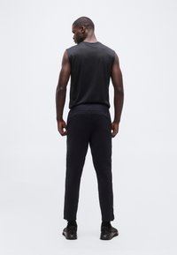 Άνδρας που στέκ εται με την πλάτη γυρισμένη, φοράει μαύρο μπλουζάκι χωρίς μανίκια, μαύρο παντελόνι και μαύρα αθλητικά παπούτσια, μπροστά σε απλό λευκό φόντο.