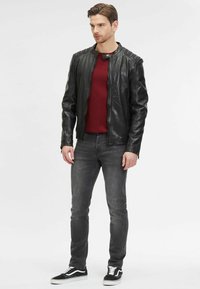 Giacca di pelle nera con colletto alto, caratterizzata da accenti texturizzati sulle spalle. Indossata sopra un maglione bordeaux e jeans grigi, abbinata a sneakers nere.