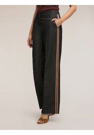 Donna che indossa pantaloni neri a gamba larga con strisce laterali marroni, top marrone senza maniche e tacchi aperti marroni, in piedi con una mano in tasca.