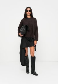Maglione a maglia marrone e peloso, gonna nera asimmetrica e stivali alti neri in pelle. Gli accessori includono una borsa nera e occhiali da sole scuri.