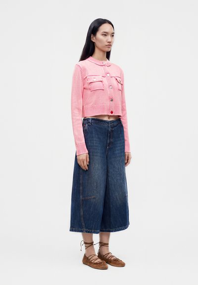 Femme portant un cardigan court rose, des culottes en denim larges et des chaussures plates marron à lacets, debout sur un fond blanc uni.