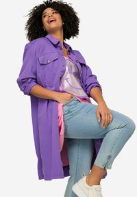 Paarse button-up jas over een roze graphic t-shirt met een glinsterend glimlachend gezicht, gecombineerd met lichtblauwe jeans en witte sneakers.