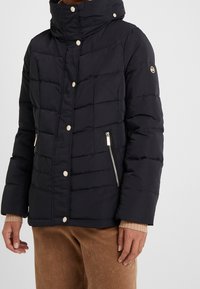 Mørk polstret jakke med høj krave. Har sølv lynlåse og knapper. Struktureret stof med et quiltet udseende, to sidelommer.