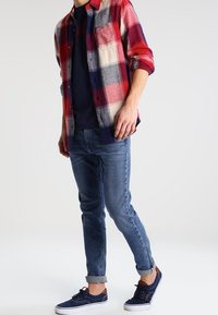 Rot- und blaukariertes Flanellhemd, navyfarbiges Rundhals-T-Shirt, hellblaue Jeans und dunkelblaue Sneakers mit Schnürsenkeln und aufgerollten Säumen.