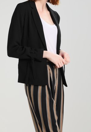 Person trägt einen schwarzen Blazer mit gebundenen Manschetten, weißes Hemd und eine hoch taillierte Hose mit vertikalen schwarzen, beigen und weißen Streifen.
