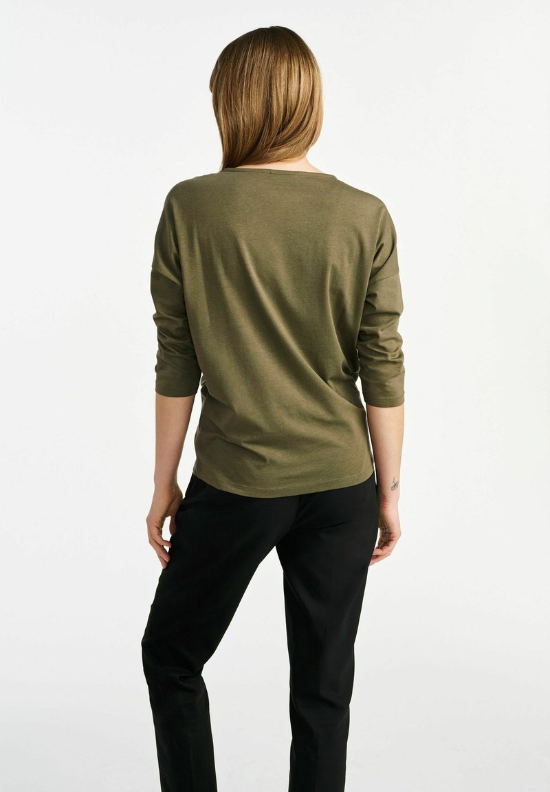 Olijfgroene langemouwtop met een relaxte pasvorm, ronde hals en zachte textuur, gecombineerd met zwarte broek, van achteraf weergegeven.