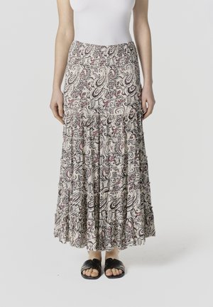 Žena nosi dlhú, viacvrstvovú sukňu s čiernymi, bielymi a červenými paisley kvetinovými vzormi, v kombinácii s čiernymi sandálmi s otvorenou špičkou.