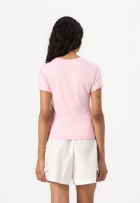 Tricou din bumbac de culoare roz deschis, cu mânecă scurtă și croială ajustată. Prezintă un decolteu rotund și o textură netedă, purtat cu pantaloni scurți albi.